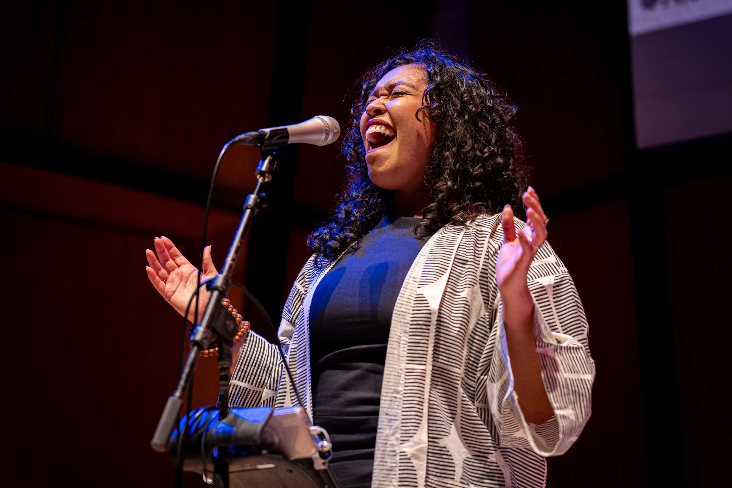 Aisha Fukushima, a woman with curly hair passionately sings into a microphone on stage, wearing a black shirt and a light patterned jacket, with her eyes closed and hands raised.
