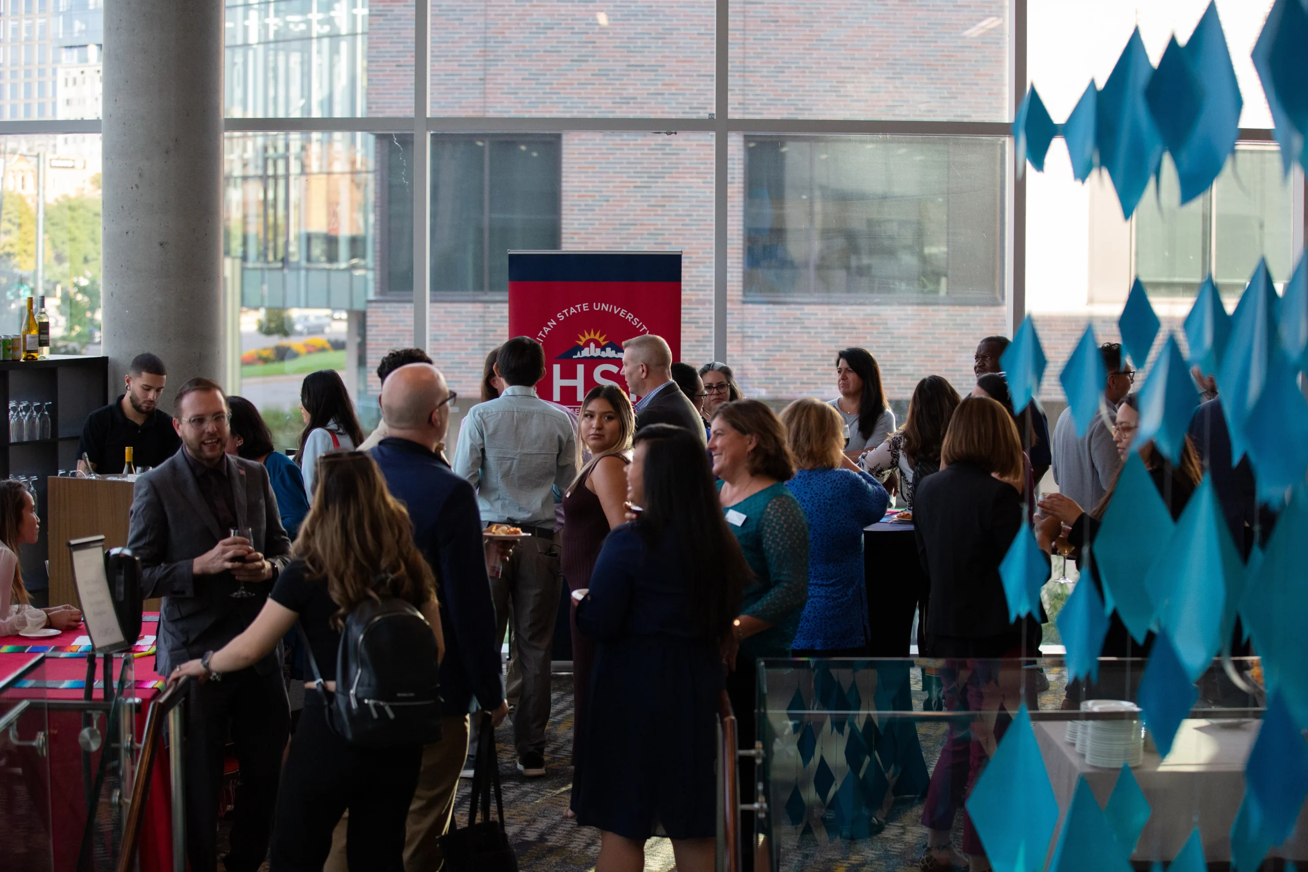 A group of people socialize and network in a bright indoor space with large windows. A red banner with HS is visible in the background and blue decorations hang to the right.